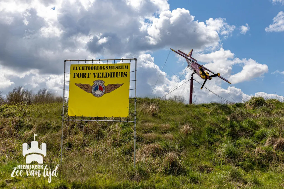 Bord met "Fort Veldhuis" erop, daarachter een klein vliegtuig op heuveltop.