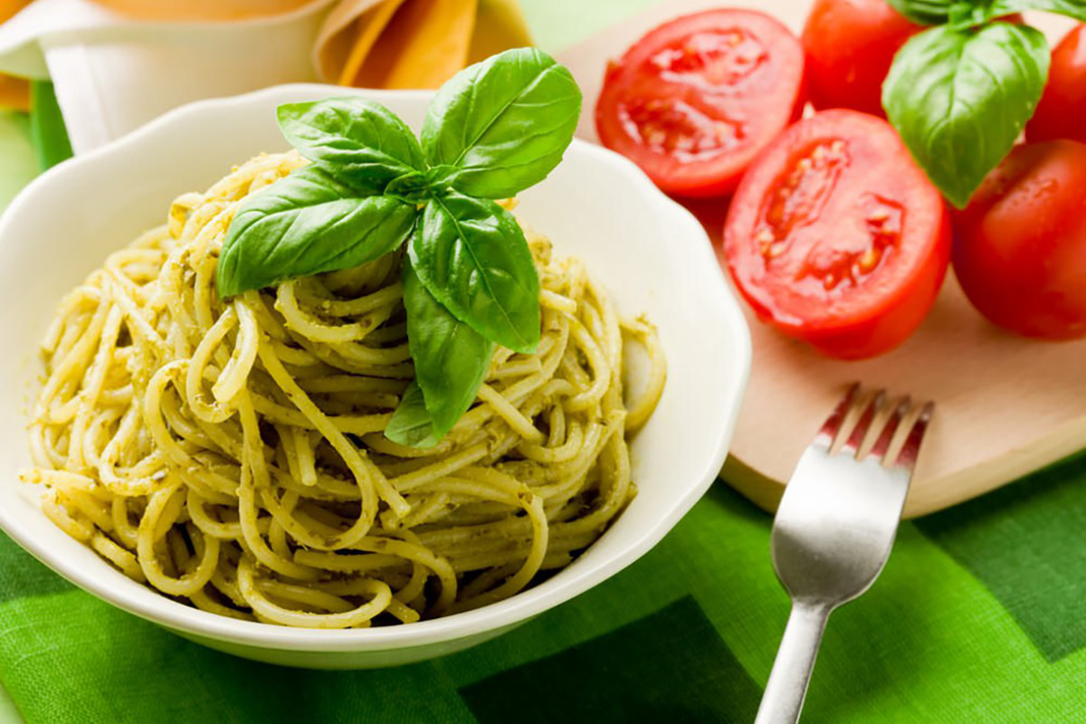 Bord paste pesto gegarneerd met basilicum en tomaten op de achtergrond.