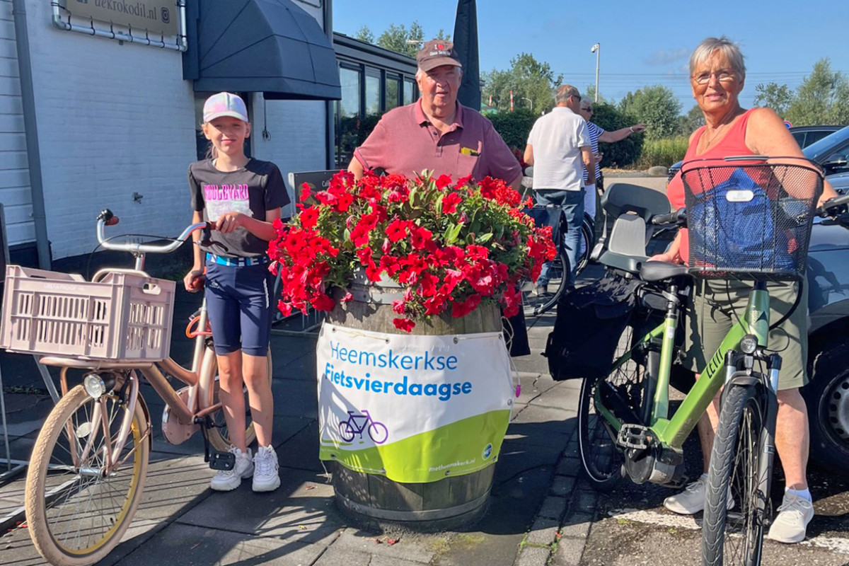 3 personen staan met de fiets aan de hand achter een plantenbak met rode bloemen