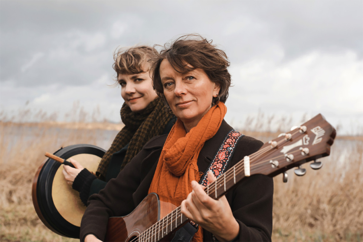 Twee vrouwen staan in de natuur met een gitaar